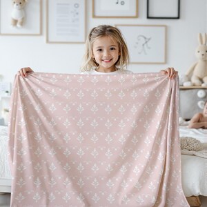 Pode incluir: Um cobertor rosa e branco com um padr&atilde;o floral branco. Uma menina est&aacute; segurando o cobertor na frente dela.