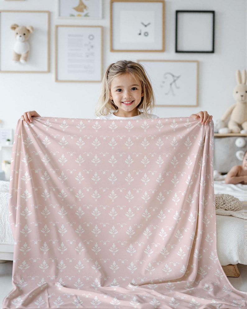 Pode incluir: Um cobertor rosa e branco com um padr&atilde;o floral branco. Uma menina est&aacute; segurando o cobertor na frente dela.