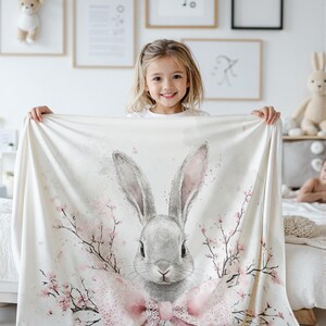 Pode incluir: Um coelho branco e rosa com um la&ccedil;o rosa e galhos de cerejeira em flor sobre um fundo branco. O coelho est&aacute; olhando para a c&acirc;mera.