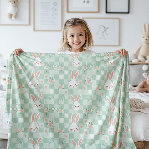 Pode incluir: Um cobertor de beb&ecirc; xadrez verde e branco com um padr&atilde;o repetido de flores rosa e rostos de coelhos brancos. O cobertor est&aacute; sendo segurado por uma menina.