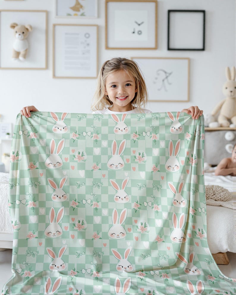 Pode incluir: Um cobertor de beb&ecirc; xadrez verde e branco com um padr&atilde;o repetido de flores rosa e rostos de coelhos brancos. O cobertor est&aacute; sendo segurado por uma menina.
