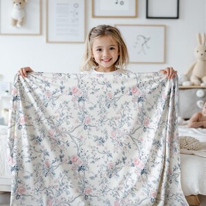 Pode incluir: Um tecido branco com estampa floral em rosa e azul. Uma menina est&aacute; segurando o tecido na frente dela.