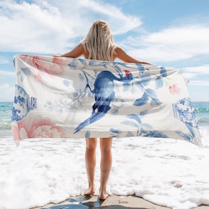 May include: A woman on a beach holding a white beach towel with a blue bird and floral design. The towel has a blue and white porcelain vase design on the corners.