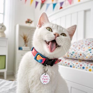 May include: A white cat with a pink and blue patterned collar and a round tag that says "Your Design Here" in pink lettering. The cat is sticking its tongue out and has a playful expression.