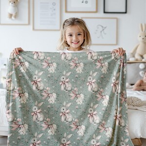 Pode incluir: Um cobertor macio e verde para beb&ecirc; com um padr&atilde;o repetido de coelhos brancos com la&ccedil;os rosa. Os coelhos s&atilde;o cercados por flores brancas e folhas verdes.