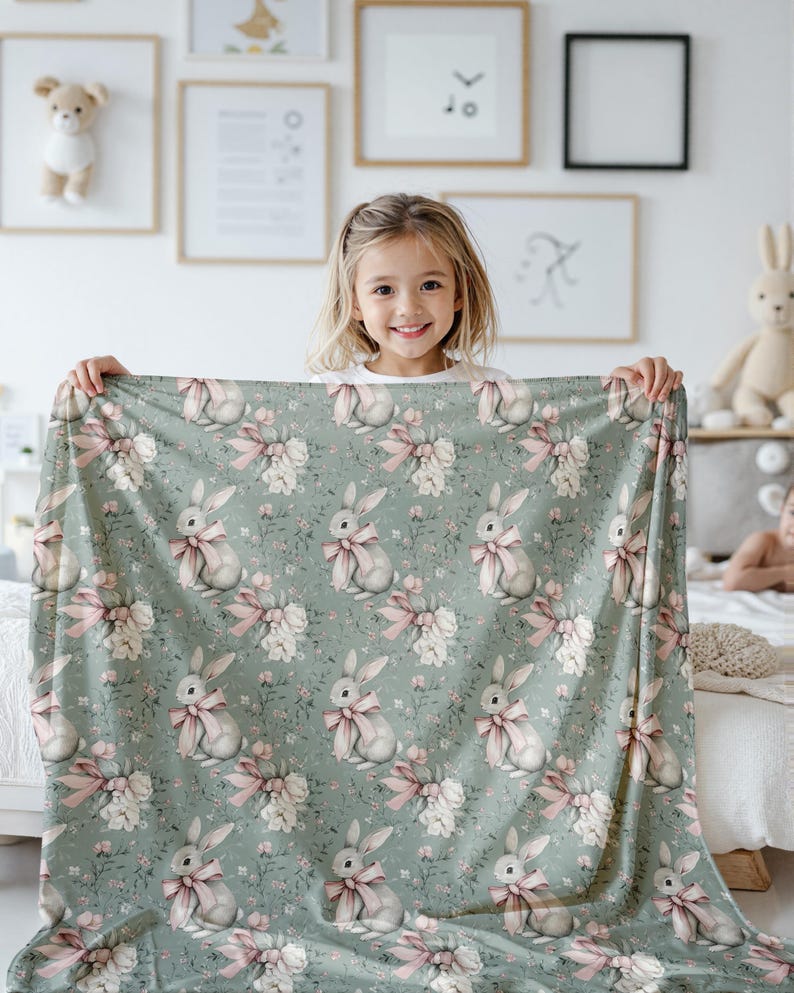 Pode incluir: Um cobertor macio e verde para beb&ecirc; com um padr&atilde;o repetido de coelhos brancos com la&ccedil;os rosa. Os coelhos s&atilde;o cercados por flores brancas e folhas verdes.