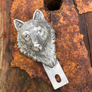 May include: A silver wolf head bottle opener with a detailed design. The opener is attached to a metal blade with a hole in the center.