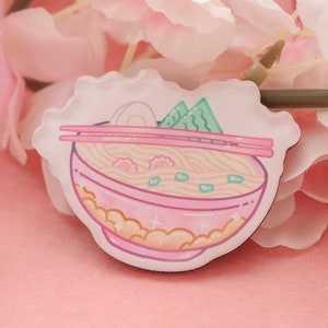 Könnte beinhalten: Eine rosa und weiße Emaille-Anstecknadel mit einer Cartoon-Illustration einer Schüssel Ramen mit Essstäbchen, einem gekochten Ei und Frühlingszwiebeln.
