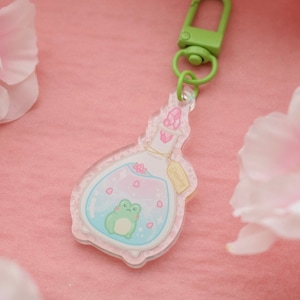 Könnte beinhalten: Ein klarer Glasflaschen-Anhänger mit einem grünen Frosch im Inneren, rosa und blauer Flüssigkeit und rosa Blütenblättern. Die Flasche hat einen Korkenverschluss und ein weißes Etikett mit der Aufschrift "Charming". Der Anhänger ist an einem grünen Metallclip befestigt.