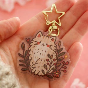 Könnte beinhalten: Ein Schlüsselanhänger aus klarem Acryl mit einer Illustration einer flauschigen beigen Katze, umgeben von dunkelbraunem Blattwerk und kleinen roten Beeren. Der Anhänger ist an einem goldfarbenen, sternförmigen Metallverschluss befestigt.