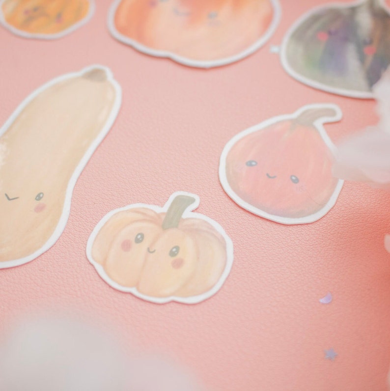 May include: A collection of pumpkin-themed stickers with smiling faces. The stickers are in various shapes and shades of orange, with green stems and white outlines. The stickers are arranged on a pink surface.
