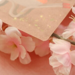 May include: A close-up of a pink and white floral arrangement with a light brown card with gold flecks on top.