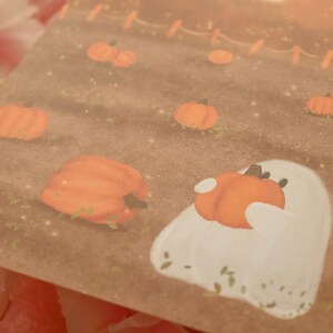 May include: A white ghost with a friendly expression holds a small orange pumpkin in its arms. The ghost is surrounded by a field of orange pumpkins on a brown background.