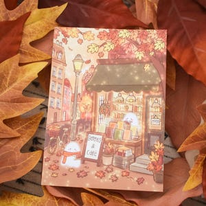 Könnte beinhalten: Ein rechteckiger Kunstdruck mit einer gemütlichen herbstlichen Straßenszene mit Buchhandlung und Café. Die Illustration zeigt zwei kleine weiße Geisterfiguren, eine davon mit orangefarbenem Schal, neben einem Schild mit der Aufschrift Books & Cafe. Warmes Licht leuchtet aus den Schaufenstern, umgeben von fallenden orangefarbenen und gelben Ahornblättern.