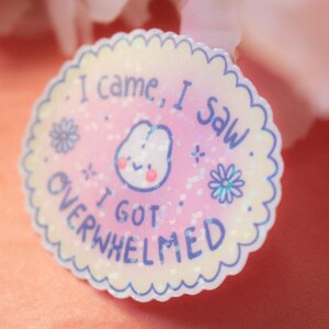 May include: A round sticker with a scalloped edge. The sticker has a pink background with the text "I came, I saw, I got overwhelmed" in blue. A cartoon character with a smiling face is in the center.