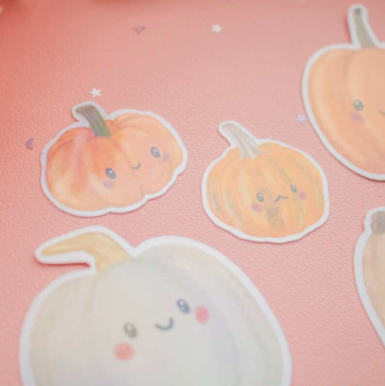 May include: A collection of pumpkin stickers with smiling faces. The stickers are in shades of orange and white, with green stems and pink cheeks. The stickers are on a pink background.