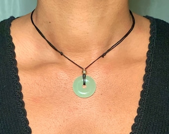 May include: A light green circular stone pendant with a central hole, suspended from a thin black cord. The pendant features a small metallic bead at the top where the cord passes through, creating a simple necklace design with a smooth finish.