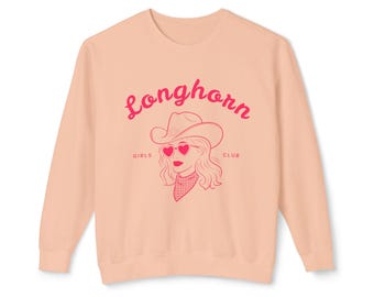 Pode incluir: Camisola de cor pêssego com um gráfico rosa de uma mulher usando um chapéu de caubói e óculos de sol em forma de coração. O texto "Longhorn Girls Club" está impresso na camisola.