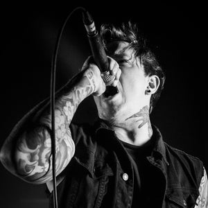 Könnte beinhalten: Ein Schwarzweißfoto eines Mannes, der in ein Mikrofon singt. Er trägt eine schwarze Weste und hat Tätowierungen auf seinen Armen und seinem Hals.