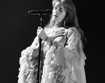 May include: A black and white image of a person singing into a microphone. The person is wearing a long, flowing dress with ruffled details and lace trim. The dress has a high neckline and long sleeves. The person is holding the microphone with one hand and has the other hand on their chest.