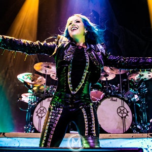 Könnte beinhalten: Eine Frau mit blauen Haaren singt in ein Mikrofon während sie auf der Bühne auftritt. Sie trägt eine schwarze Lederjacke mit silbernen Nieten und eine schwarze Hose mit silbernen Akzenten. Die Bühne ist mit bunten Lichtern beleuchtet und hinter ihr steht ein Schlagzeug.