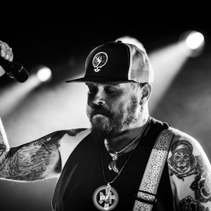 Könnte beinhalten: Ein Schwarzweißfoto eines Mannes mit einer schwarzen Mesh-Baseballkappe mit weißem Logo, einem schwarzen Tanktop und einem Gitarrengurt. Er hat einen Bart und Tattoos auf den Armen.