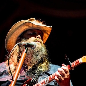 May include: A musician with a long, gray beard and a cowboy hat plays a guitar. The guitar has a wooden neck and a red body. A microphone is positioned in front of the musician. The background is dark.