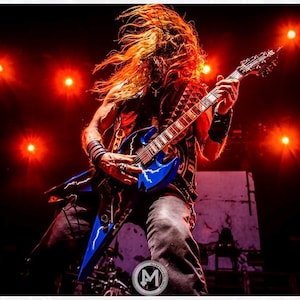 May include: A musician with long, flowing hair plays a blue electric guitar on stage. The guitar has a lightning bolt design. The stage is lit with red lights, creating a dramatic atmosphere. The musician is wearing a vest and wristbands.