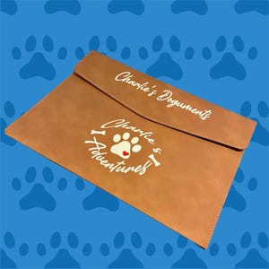 May include: A brown leather folder with a white paw print and bone design. The text "Charlie's Documents" and "Charlie & I Adventures" is printed on the folder.