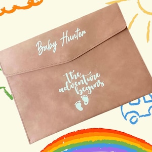 May include: A light pink leather folder with the text "Baby Hunter" and "The adventure begins" in white lettering. There are two small white footprints below the text.
