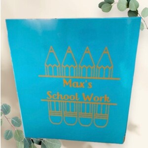 May include: A turquoise blue file folder with gold lettering that says "Max's School Work". The folder has a design of three gold pencils on the top and five gold pencils on the bottom.