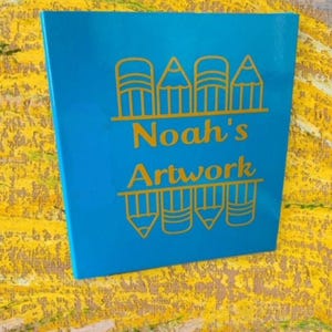 May include: A blue sign with gold lettering that says "Noah's Artwork" with a row of gold pencils above and below the text.
