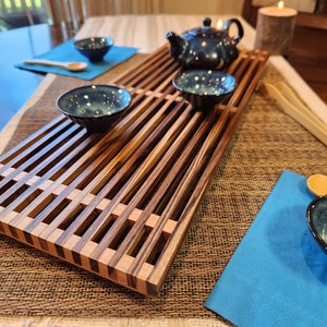 May include: A wooden tea tray with a slatted design, featuring four blue and white teacups and a blue teapot. The tray is set on a woven mat with a blue napkin and a wooden spoon.