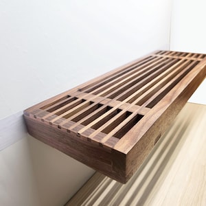 May include: A rectangular wall-mounted shelf crafted from dark brown wood, featuring a slatted top surface with evenly spaced vertical openings. The shelf has a solid frame and a smooth finish, showcasing the natural grain patterns of the wood.