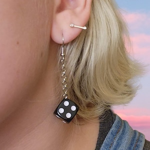 May include: A close-up of a person's ear with two earrings. One is a small silver bar stud. The other is a dangling earring with a black die with white dots attached to a silver chain.