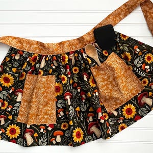 K&ouml;nnte beinhalten: Schwarze Sch&uuml;rze mit Sonnenblumen- und Pilzmuster, mit zwei Taschen und passendem Bund. Ein Holzl&ouml;ffel und ein schwarzer Spatel liegen auf der Sch&uuml;rze. Die Sch&uuml;rze ist aus Stoff.