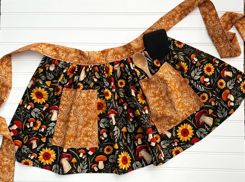K&ouml;nnte beinhalten: Schwarze Sch&uuml;rze mit Sonnenblumen- und Pilzmuster, mit zwei Taschen und passendem Bund. Ein Holzl&ouml;ffel und ein schwarzer Spatel liegen auf der Sch&uuml;rze. Die Sch&uuml;rze ist aus Stoff.