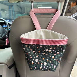 May include: A floral patterned fabric car trash can with a pink and white gingham trim hangs from the headrest of a car seat.
