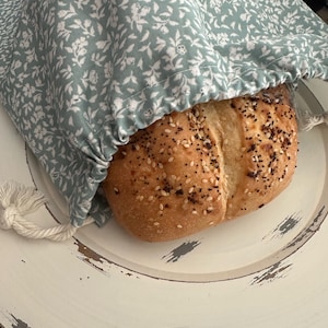 May include: A loaf of bread with sesame seeds peeking out from a light green floral drawstring bread bag. The bag is on a distressed white plate.