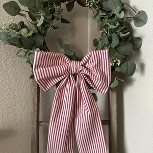 May include: A decorative wreath featuring a circular base adorned with green foliage and small white flowers. A large bow made of red and white striped fabric is centered on the wreath. The wreath is hanging on a wooden ladder.