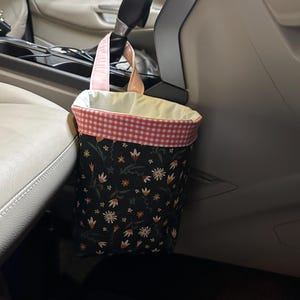 May include: A black and pink floral patterned trash can with a pink gingham trim hangs from the headrest of a car seat.