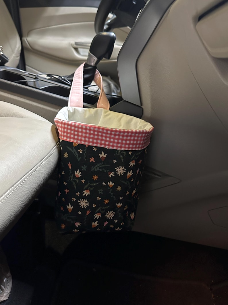 May include: A black and pink floral patterned trash can with a pink gingham trim hangs from the headrest of a car seat.