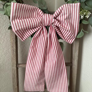 May include: A large decorative bow made of red and white striped fabric. The bow is attached to a wooden ladder and surrounded by greenery. The bow's fabric has a classic ticking stripe pattern, perfect for farmhouse or country decor.