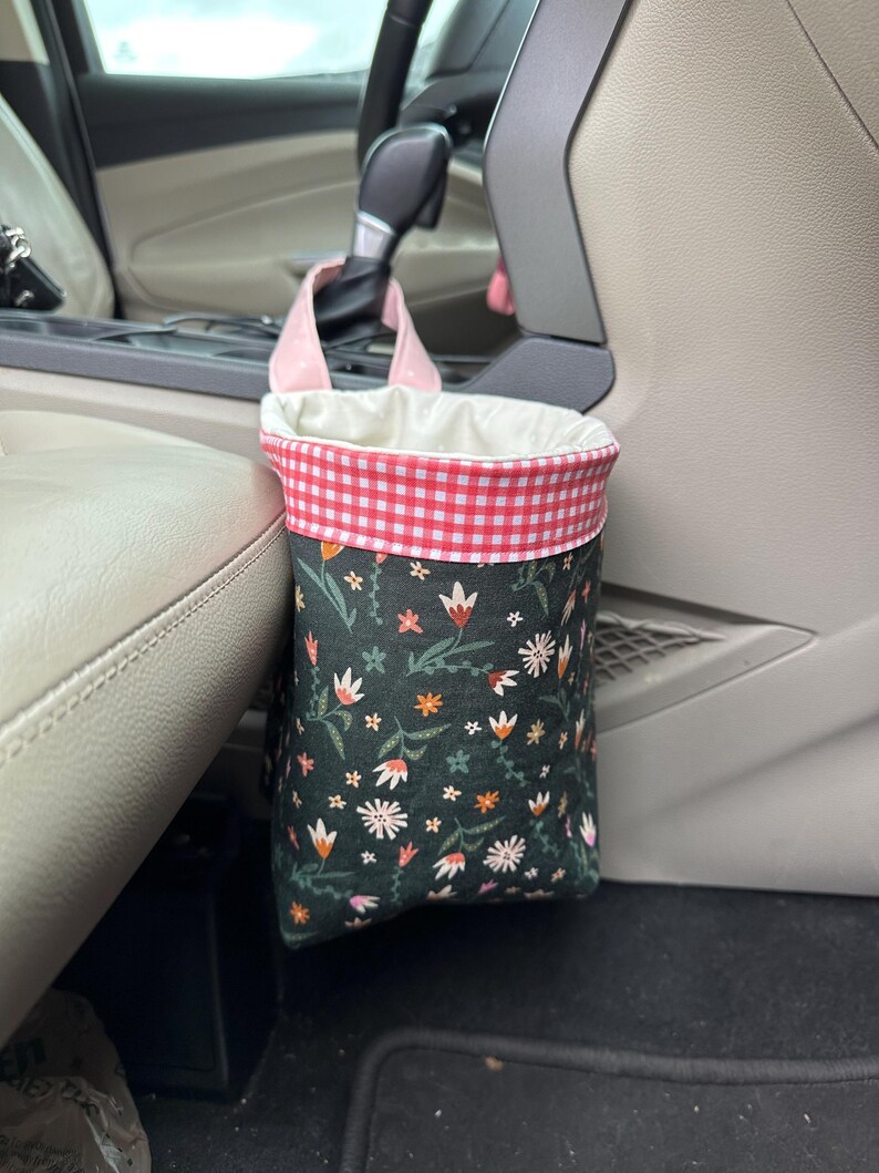 May include: A floral patterned fabric trash can with a pink and white gingham trim hangs from the headrest of a car seat.