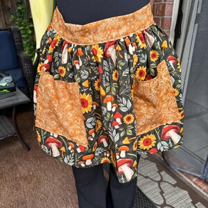 K&ouml;nnte beinhalten: Eine Halbsch&uuml;rze mit dunklem Hintergrund, mit einem Muster aus Sonnenblumen, Pilzen und Bl&auml;ttern in Orange-, Gelb- und Wei&szlig;t&ouml;nen. Die Sch&uuml;rze hat zwei Taschen und ein dekoratives Band oben.