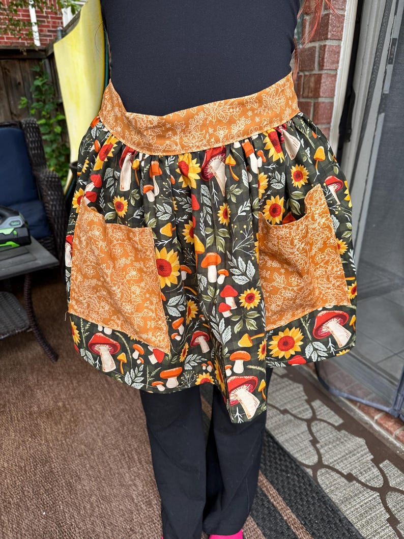 K&ouml;nnte beinhalten: Eine Halbsch&uuml;rze mit dunklem Hintergrund, mit einem Muster aus Sonnenblumen, Pilzen und Bl&auml;ttern in Orange-, Gelb- und Wei&szlig;t&ouml;nen. Die Sch&uuml;rze hat zwei Taschen und ein dekoratives Band oben.