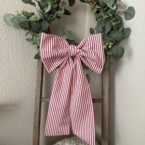 May include: A decorative wreath with green eucalyptus leaves and small white flowers. A large bow made of red and white striped fabric is attached to the wreath. The wreath is hanging on a wooden ladder.
