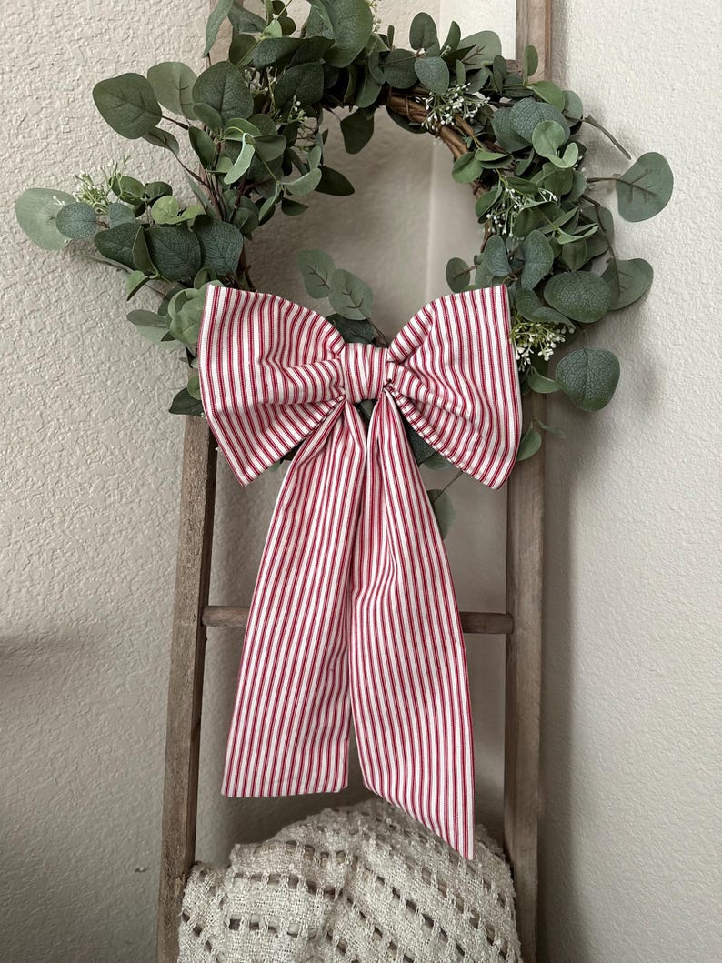 May include: A decorative wreath with green eucalyptus leaves and small white flowers. A large bow made of red and white striped fabric is attached to the wreath. The wreath is hanging on a wooden ladder.