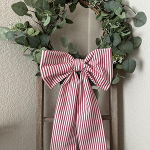 May include: A decorative wreath featuring green eucalyptus leaves and small white flowers. A large bow made of red and white striped fabric is attached to the wreath. The wreath is hanging on a wooden ladder.