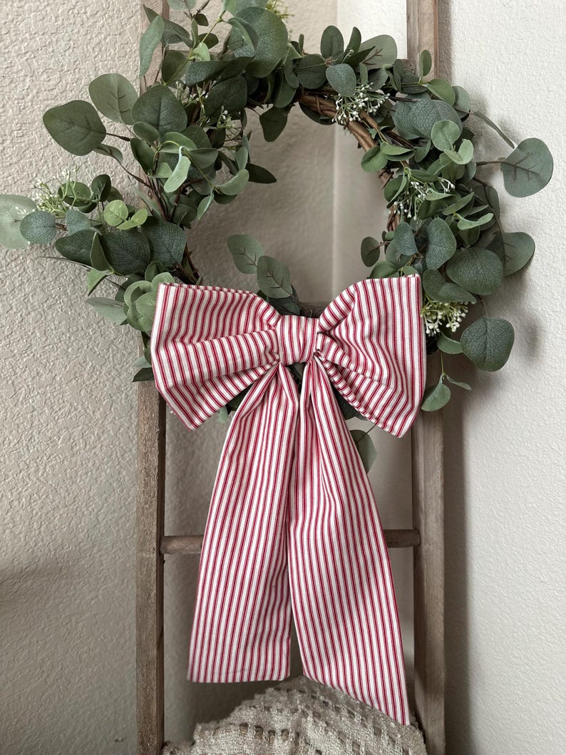 May include: A decorative wreath featuring green eucalyptus leaves and small white flowers. A large bow made of red and white striped fabric is attached to the wreath. The wreath is hanging on a wooden ladder.
